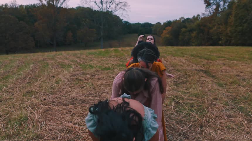 A group of women in dresses dance in a field of dry grass