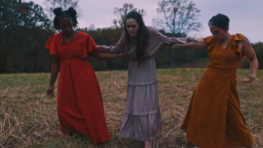 Three women in different colored dresses dancing together in a field