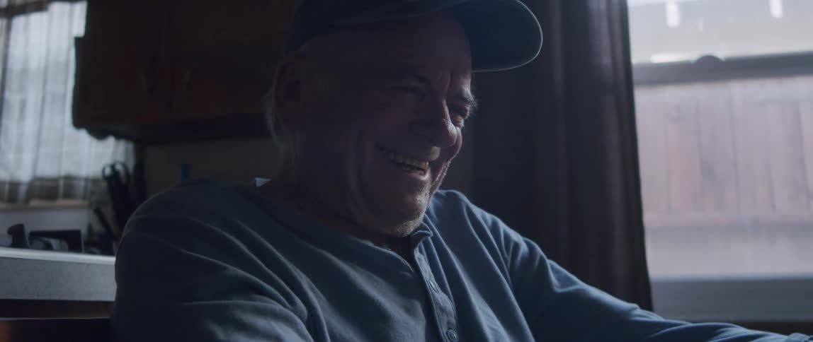An elderly man sits in his kitchen laughing as he looks at a tablet