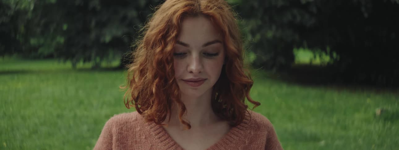 A red headed teenage girls stands in a forest on an open grassy area while the wind blows her hair