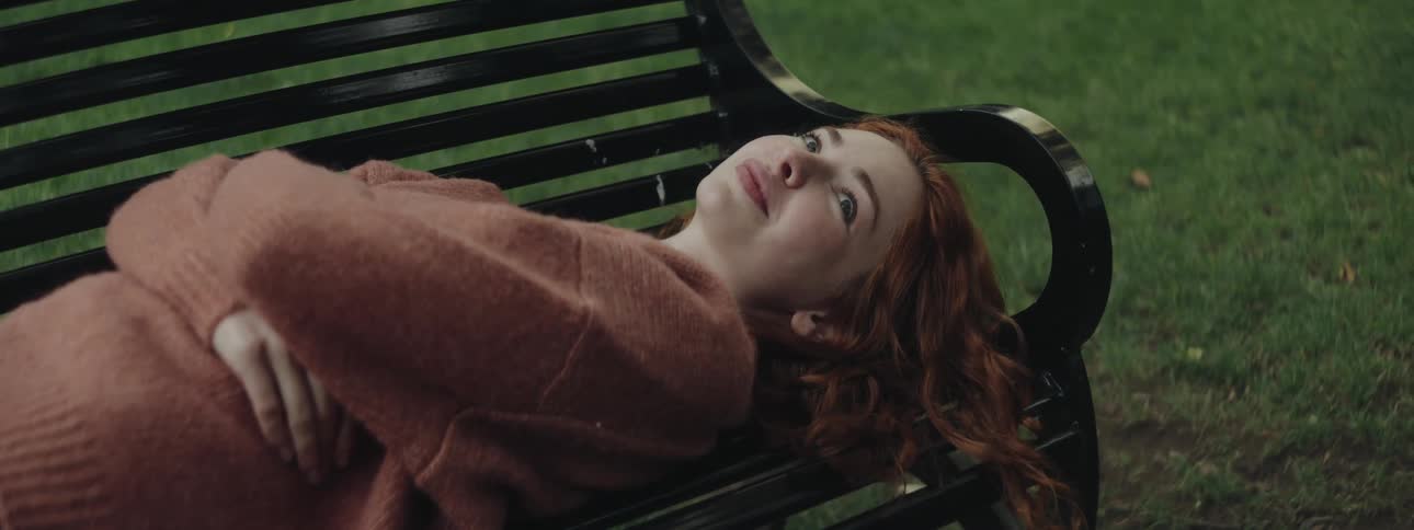 A young redhead woman in a pink sweater rests on a park bench