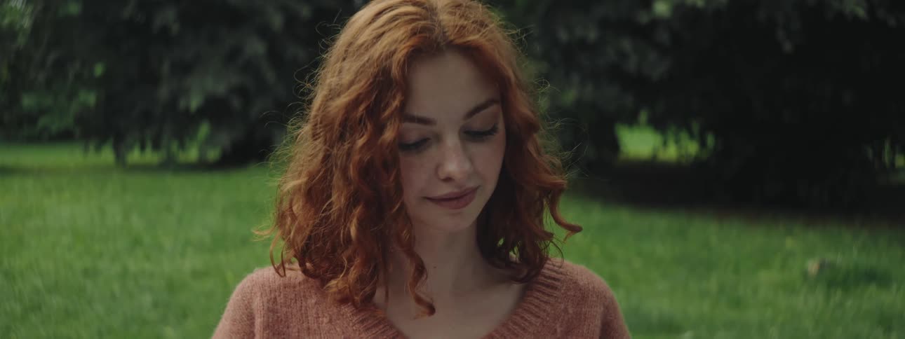 Red haired woman sitting outdoors near a grove smiles while focusing on an enjoyable activity