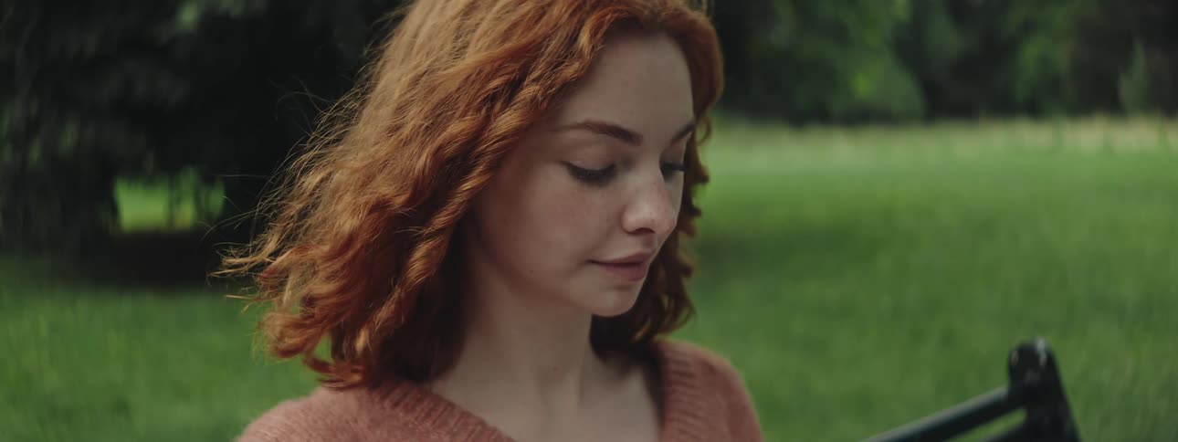 A redheaded teen girl sits on a bench on a grassy plain while reading a book