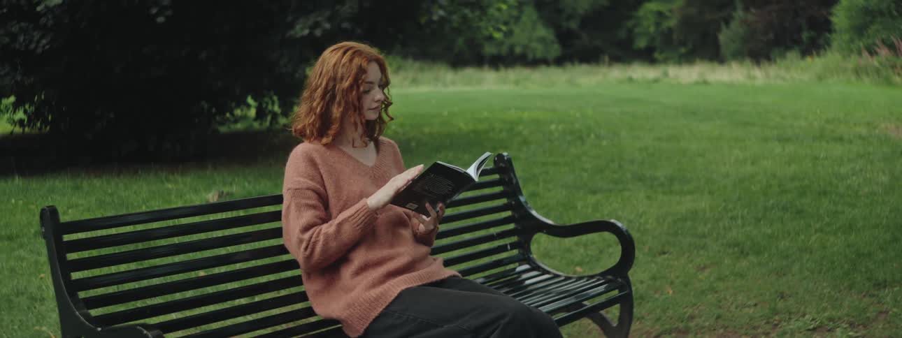 A redhead teenage girl sits on a bench on a grassland and reads a book