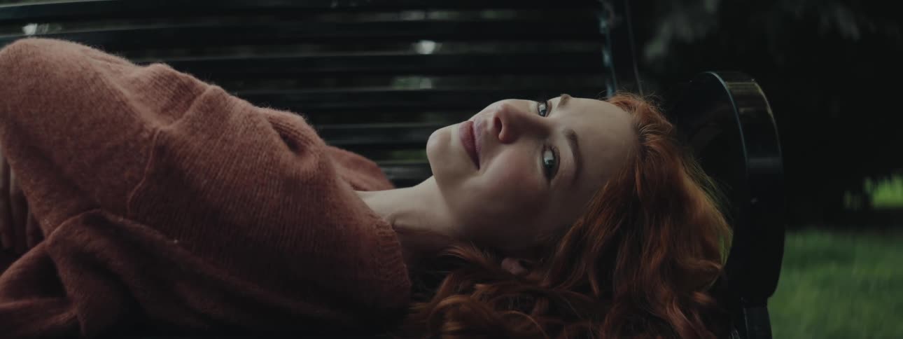 A teenage girl with red hear and wearing a sweater reclines on a bench as she turns her head
