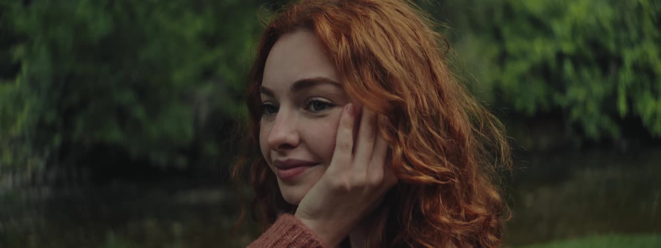 A pretty red haired teenage girl turns her head around and places her hand on her cheek as she smiles