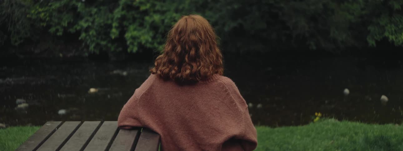 A young redhead woman sits in a park facing away from the camera