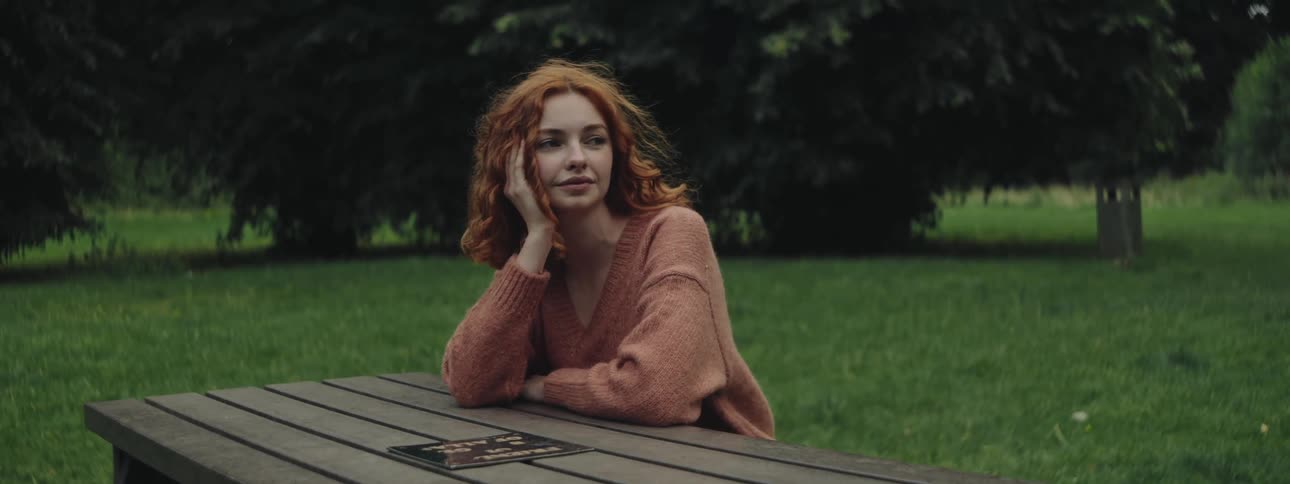 A woman with fiery red hair wearing a cozy brown sweater sits at a rustic wooden desk in a lush green park