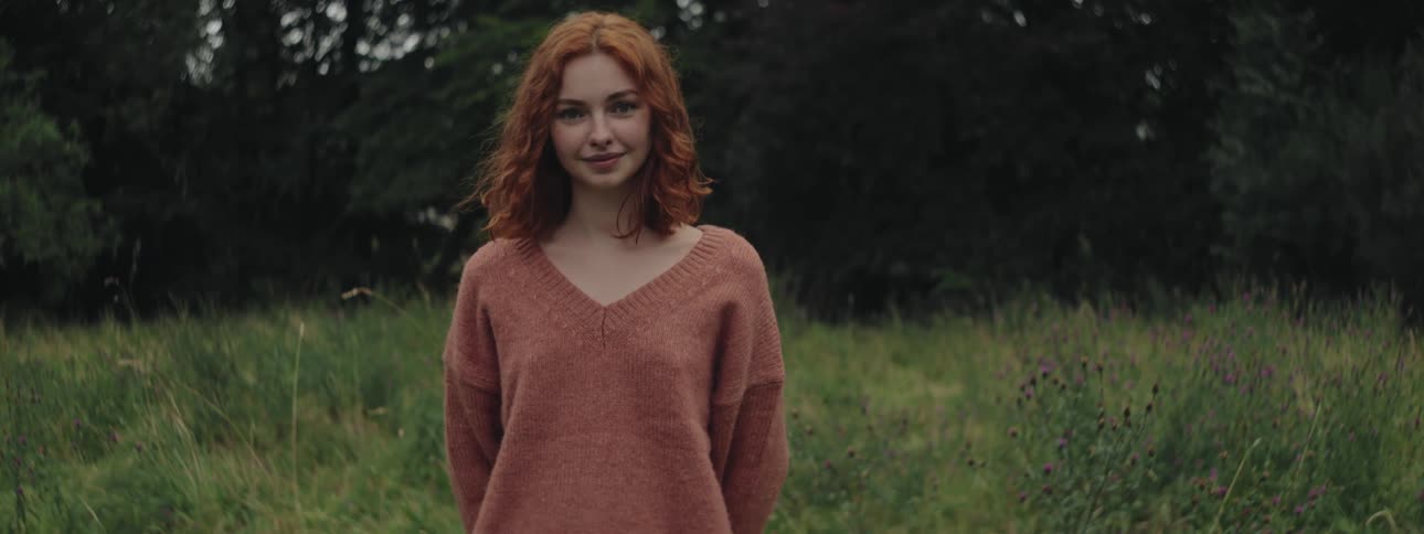 Redheaded woman smiles while posing in a field near a grove of trees