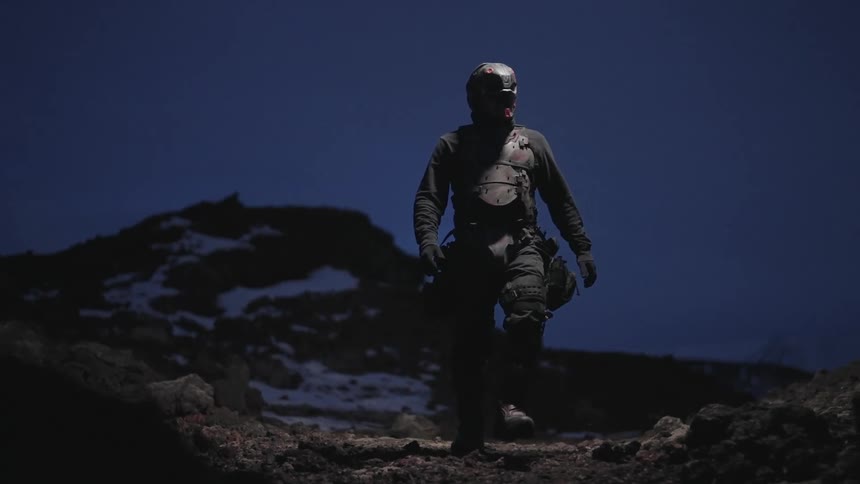 A masked figure in tactical gear walks along a snowy mountain ridge at night