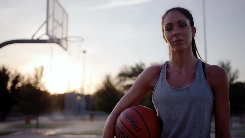 A woman in a gray tank top holding an orange basketball looking forward and behind her is an outdoor basketball court