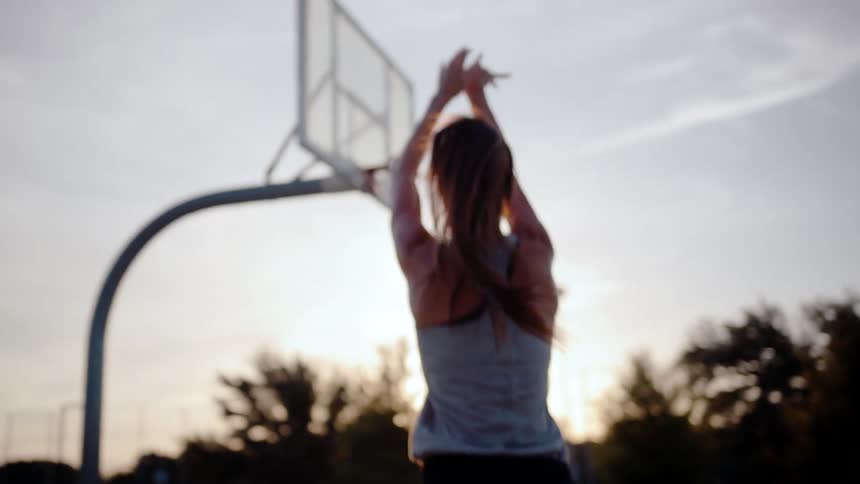 A female basketball player shoots a basketball into the hoop at dusk
