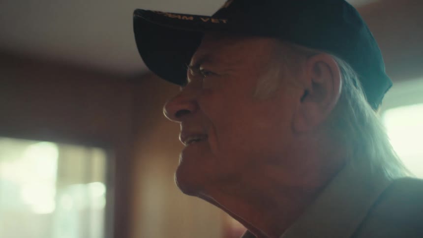 An old man stands in a living room smiling and squinting