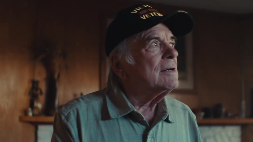 A man in a vietnam veteran hat stares off into the distance with a serious expression
