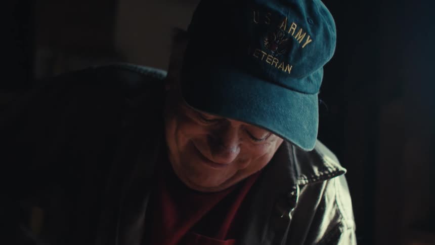 A man wearing a us army veteran hat laughs brightly in a dimly lit room