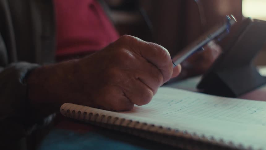 An old man writes in a notebook and then puts his glasses on
