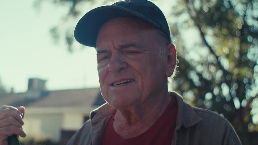 An old man in a ball cap smiles outdoors while using a hand tool