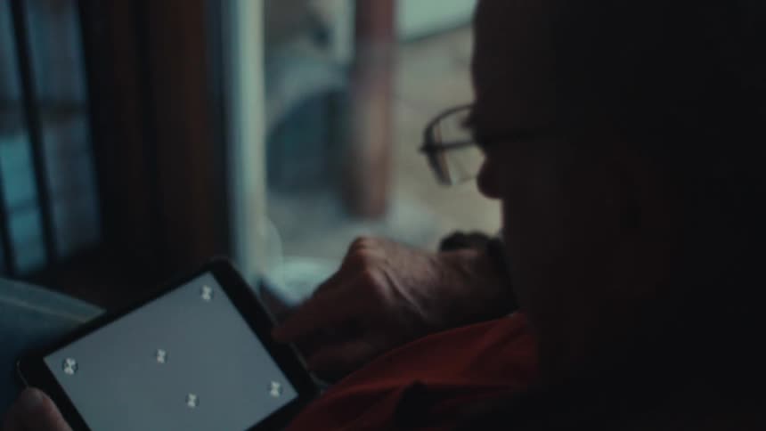 An older man in glasses is using a tablet while sitting in a chair