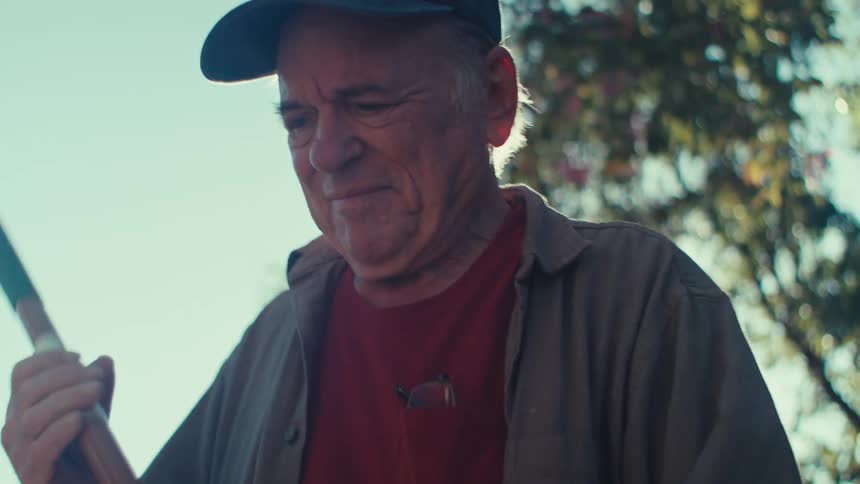 An old man smiles as he works outside in a yard with a rake