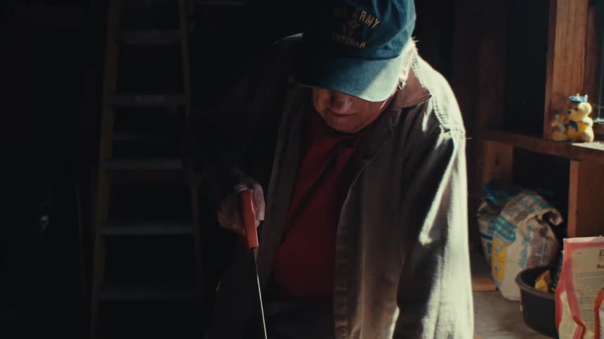 On old man saws a piece of wood in a dark workshop
