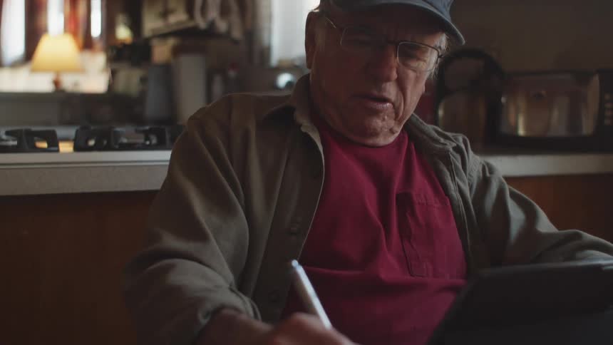 Elderly man writes while sitting at his kitchen table and using a tablet