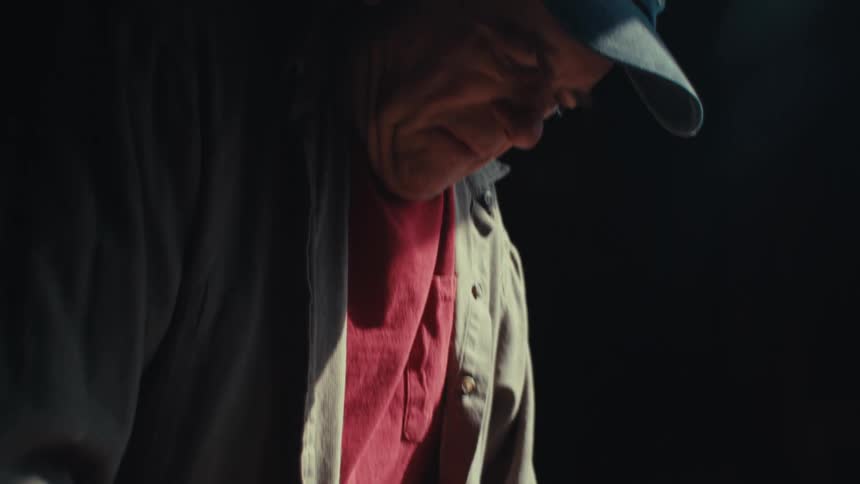 An old man measures wood in a workshop before looking up and thinking