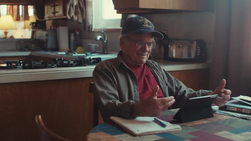 An older man wearing a us army veteran hat sits at a table in a kitchen and uses a tablet smiling and gesturing