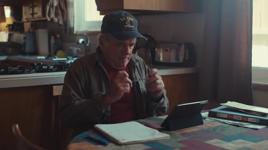A man wearing a cap enters a kitchen and then sits at a table where there is a tablet in front of him he puts on his glasses and starts using the tablet