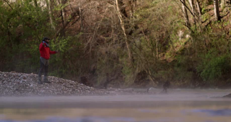A man standing on a riverbank is casting out a fishing line