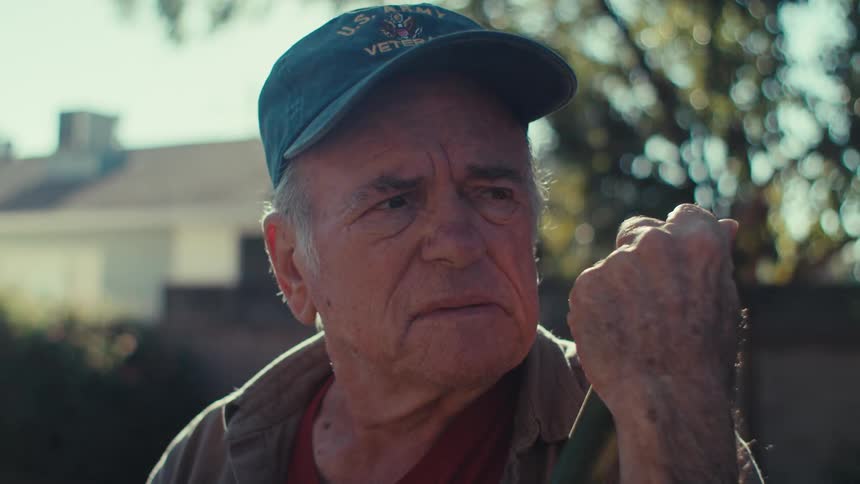 An old man leans on a rake outdoors while looking upset