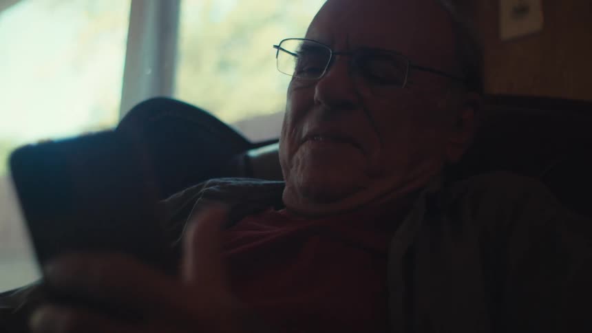 Elderly man sitting at home smiles while scrolling on a smartphone