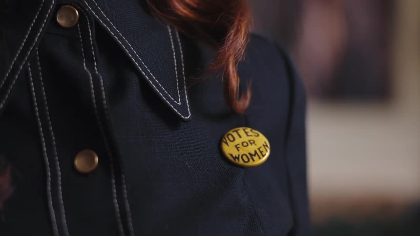 Redheaded woman pins on a button with a slogan relevant to votes for women
