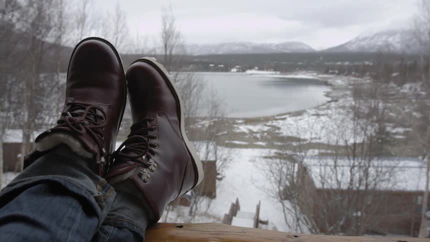 A man is resting his feet on a porch railing overlooking a snowy landscape