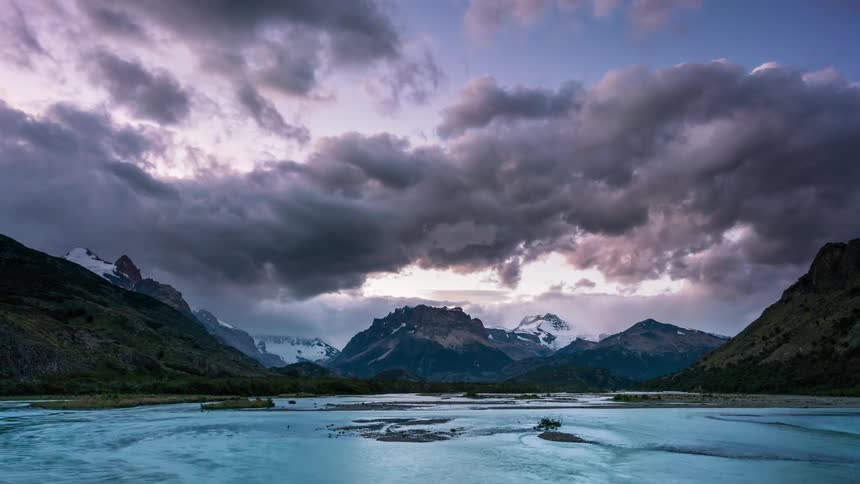 A dramatic landscape with a winding river flowing through a valley framed by snowcapped mountains and a stormy dramatic sky