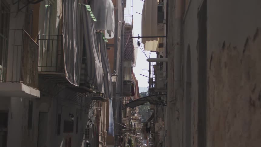 A narrow winding alleyway in a european city lined with tall buildings and balconies filled with laundry drying in the sun