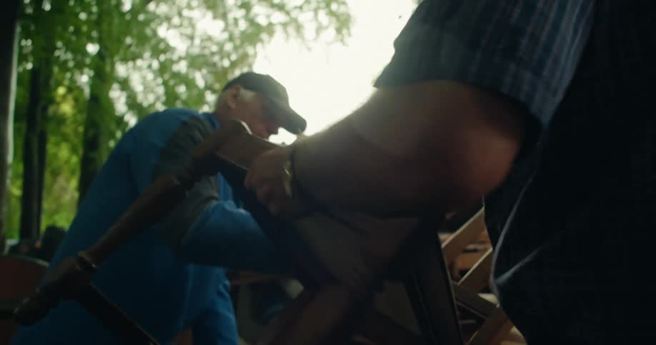 A man carries a wooden chair over to another man who loads it onto a pile outdoors