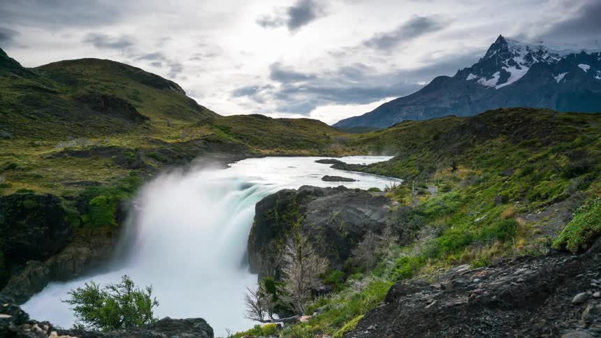 A powerful waterfall cascades down a rocky cliff with a snow capped mountain and lush vegetation in the background