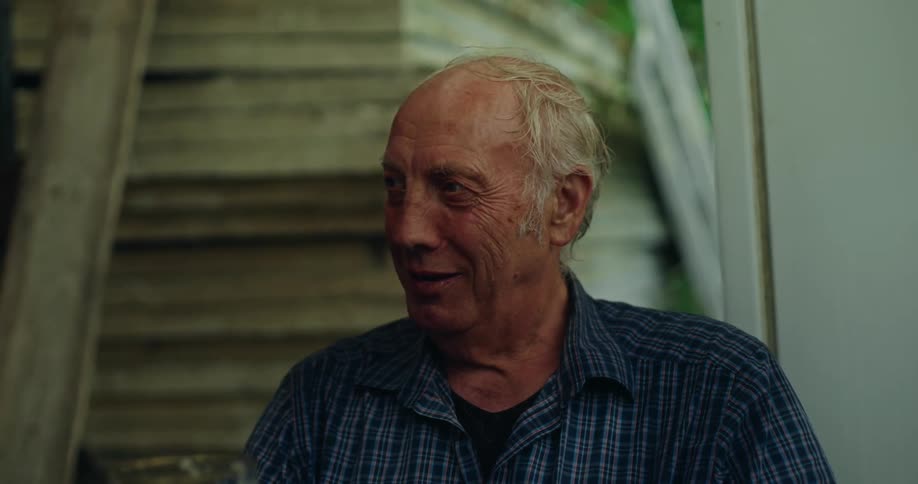 An old man stands in a building while speaking and holding a glass of beer