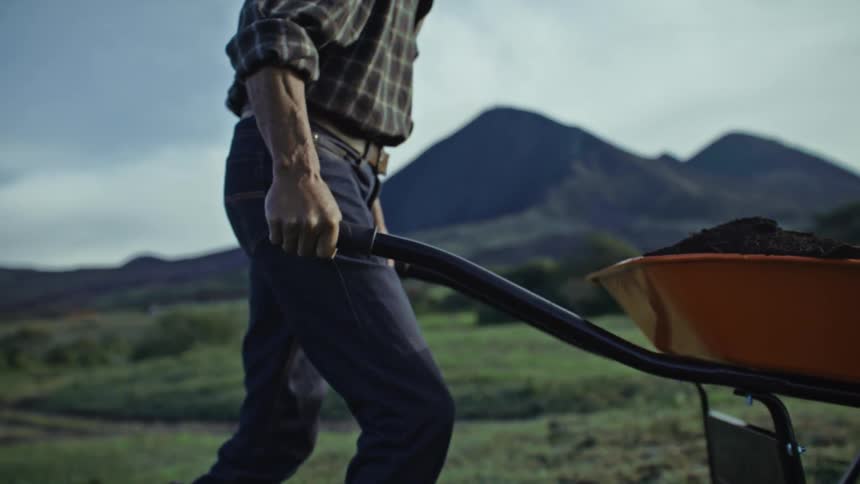 A man lifts the handles of a wheelbarrow full of dirt and pushes it across a field