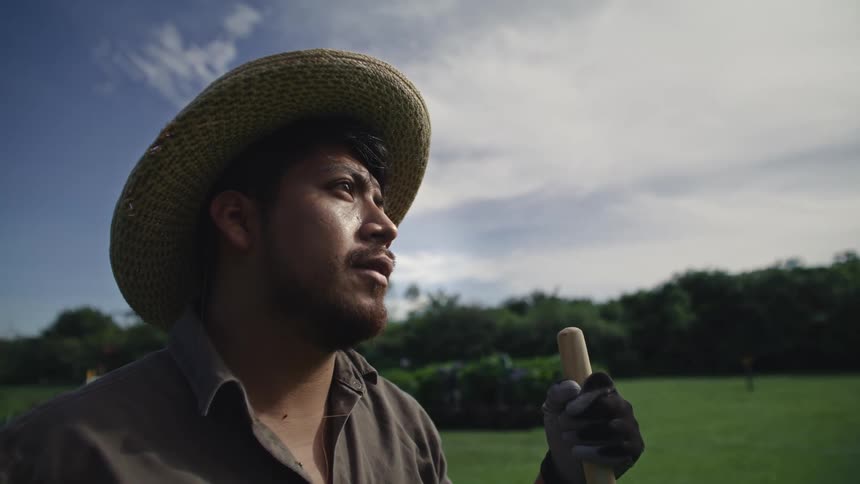 A man with a straw hat holding a tool in his hands looking forward and he is in a grass field