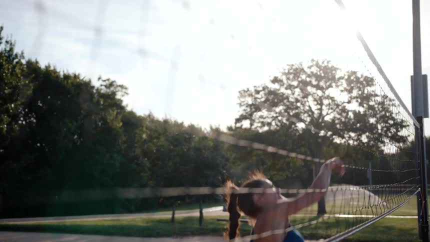 A woman wearing a blue top is jumping to hit a volleyball that is going over a net