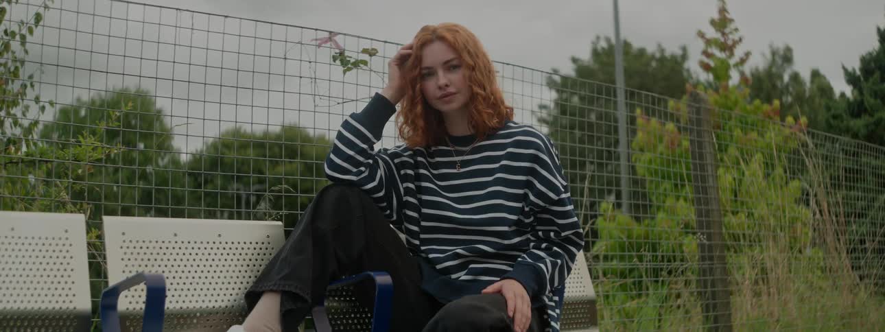 A young naked woman sitting on an outdoor bench with one hand on her chin while looking straight ahead