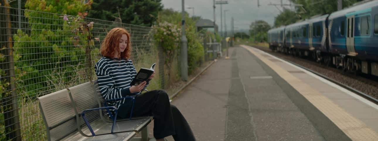 Redheaded woman sitting on a bench outdoors reads while a train passes nearby
