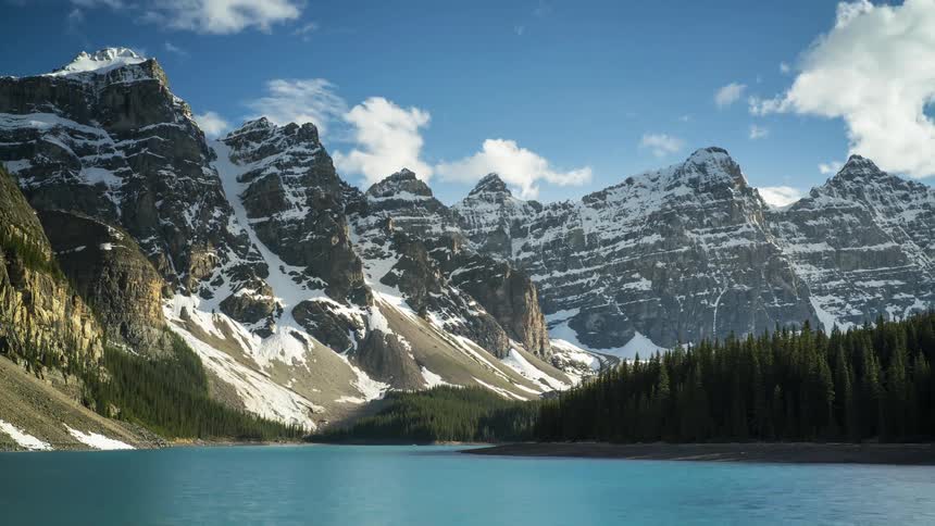 Shadows cover snowy mountains and the nearby forest and lake they tower over as a winter day passes