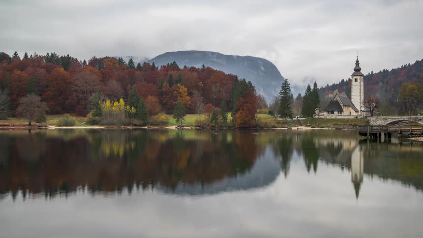 Natural landscape of a lake next to a forest and a cottage with a tower with mountains on the background during day time