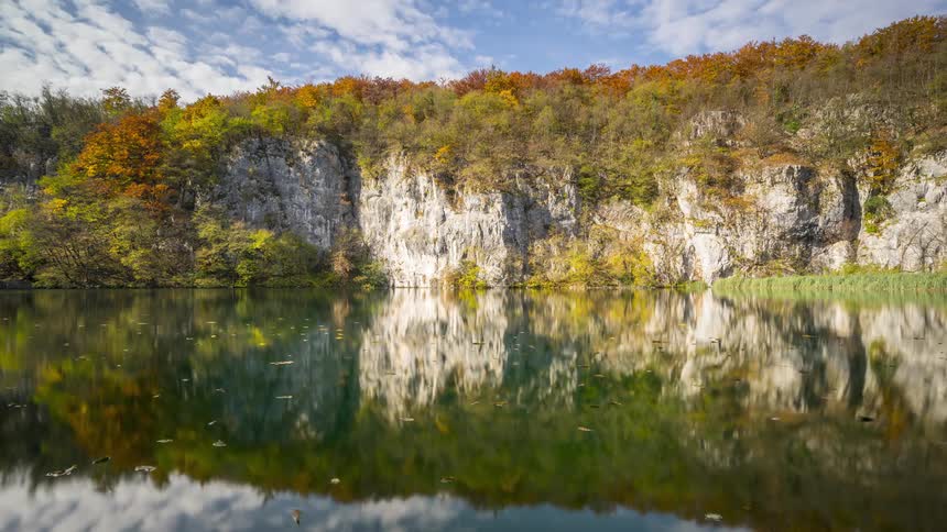 A serene lake reflects the colorful autumn trees and cliffs lining its shore on a partly cloudy day