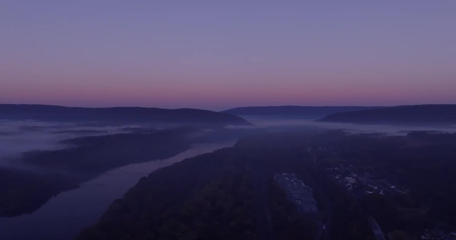 A river winds through a misty valley as the sun rises casting a soft pink glow on the horizon