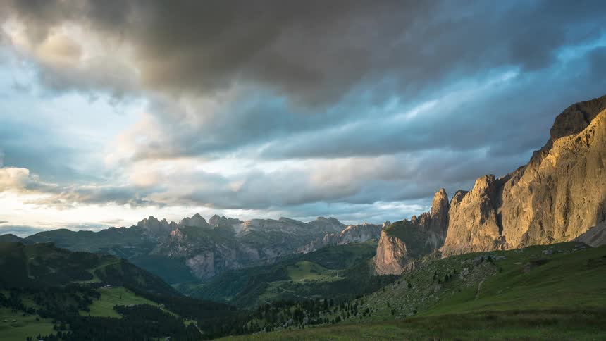 A mountainous region with large fields covered with green grass and gray clouds passing in the sky on a clear day