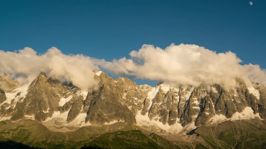 A snow capped mountain range under a vibrant blue sky with fluffy clouds showing the dramatic peaks and changing colors of sunrise