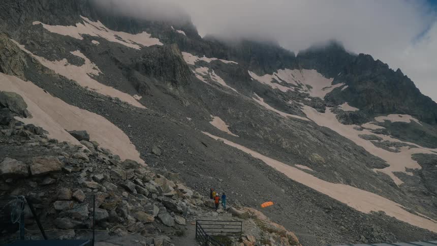 Hikers ascend a steep rocky mountainside with peaks shrouded in mist and patches of snow on the higher slopes